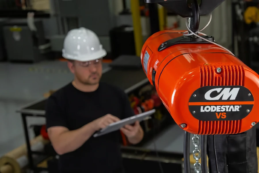 A man in a white hard hat uses a tablet near an orange CM Lodestar VS electric chain hoist in an industrial setting.