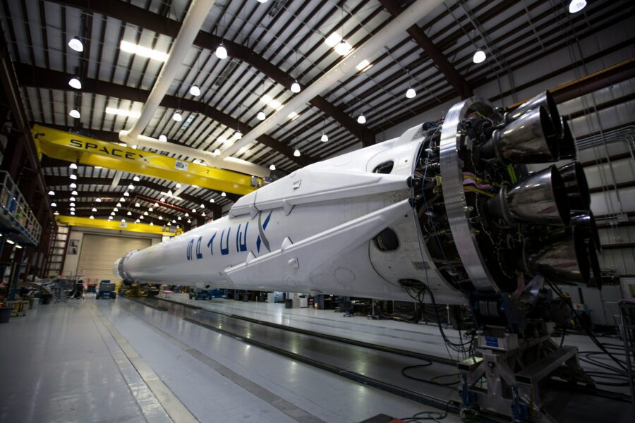 A SpaceX Falcon 9 rocket lies horizontally in an indoor hangar, showing its engine section and structural details under bright overhead lights.