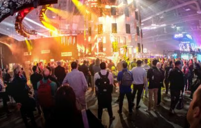 A large crowd of people stands indoors facing a brightly lit stage with colorful lights and screens during an event or concert.