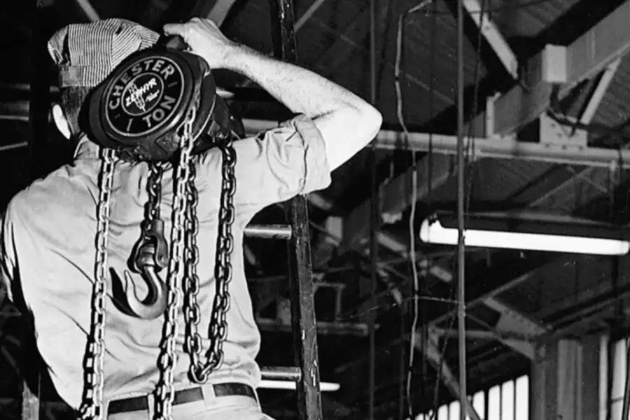 A worker in uniform stands on a ladder operating a Chester hoist inside an industrial facility with visible beams and overhead lighting.