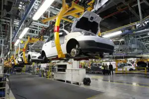 Cars on an assembly line in a factory, with partially assembled vehicles suspended and workers in the background.