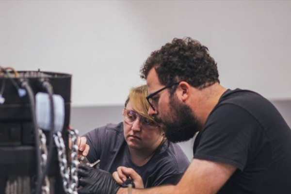 Two people wearing safety glasses work together on a mechanical project at a table, handling tools and components including chains and wires.