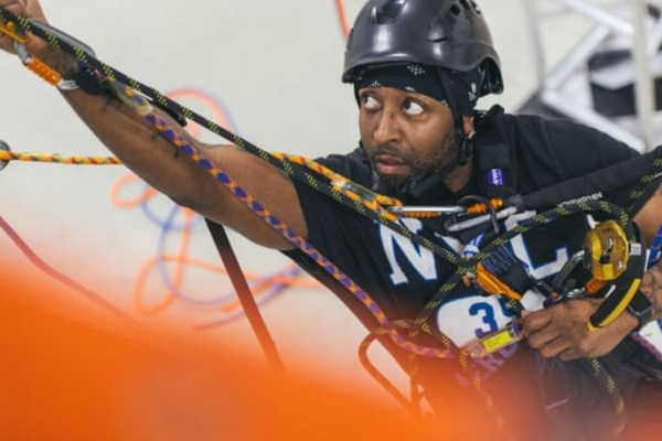 A man wearing a helmet and harness climbs using ropes and safety equipment in an indoor setting.