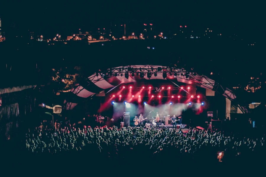 Large crowd attends an outdoor nighttime concert, with a band performing under a canopy stage lit by red and white spotlights. City lights are visible in the background.