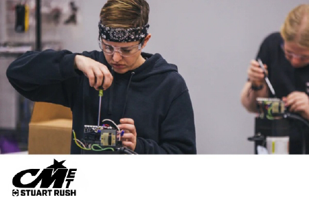 Two individuals work on small mechanical or electronic devices at a table, using tools. One wears safety glasses and a bandana. CMT Stuart Rush logo appears in the lower left corner.