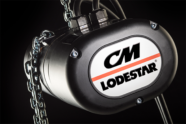 A close-up of a black CM Lodestar electric hoist with a chain and cables attached, shown against a dark background.