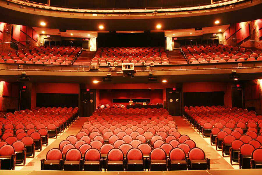 Empty theater auditorium with rows of red cushioned seats facing a stage, and a balcony level visible in the background.