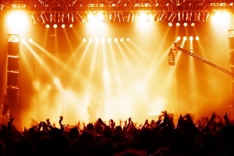 A large crowd at a concert with raised hands, bright stage lights, and heavy smoke, silhouetting performers on stage.