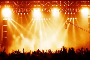 Crowd with raised hands at a concert in front of a brightly lit stage with orange and yellow lights.