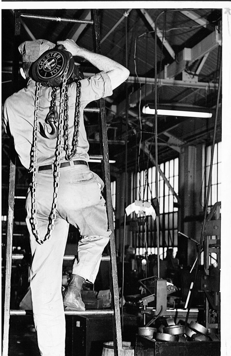 A worker in uniform stands on a ladder and lifts a heavy load using a chain hoist inside an industrial workshop with machinery and tools visible in the background.