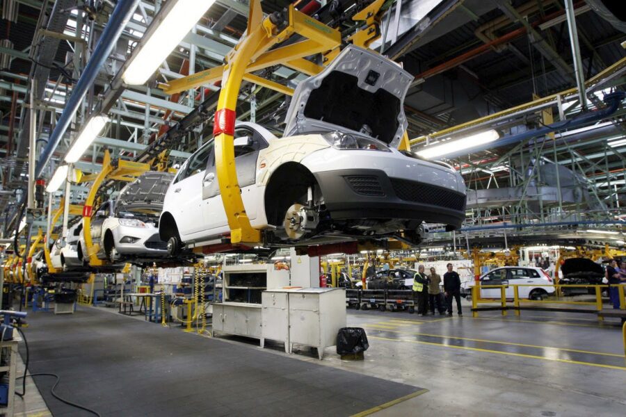 Assembly line in a car manufacturing plant with partially assembled white vehicles suspended by yellow robotic arms.