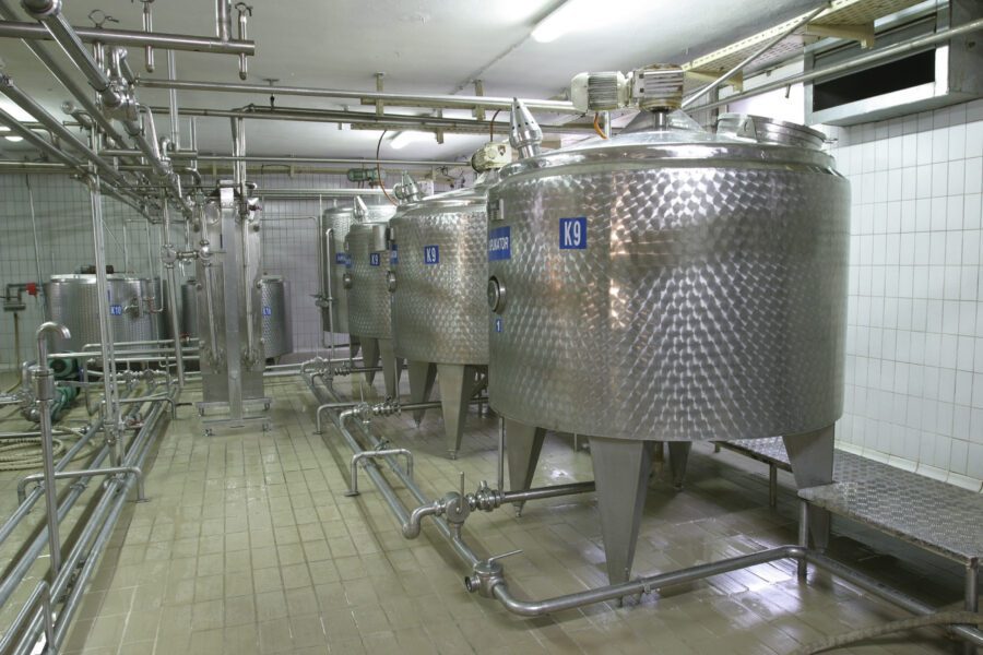 A row of large stainless steel tanks and pipes inside a clean, tiled industrial processing facility.