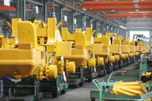 Rows of yellow heavy machinery or vehicle parts are lined up in a large industrial factory with overhead cranes.