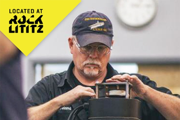 A man wearing a navy cap and glasses works on a piece of equipment; a yellow triangle with the text "Located at Rock Lititz" is in the upper left corner.